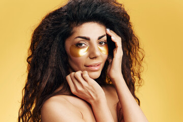 Enjoyed pretty Latin lady in gold hydrogel patches under eye touch perfect flawless skin posing isolated over pastel yellow background. Cosmetic product ad Natural beauty concept Studio portrait