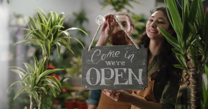 Flower shop employees hanging welcoming wooden open sign on glass entrance door. Happy employees starting new workday 4k footage