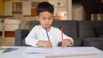 Little asian boy in white drawing using both of hands with blue and red pen on paper sitting at home