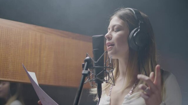 Beautiful female singer working in music studio while recording new song. Blonde Caucasian girl wearing headphones and singing song in studio while reading lyrics from paper. Music, art concept