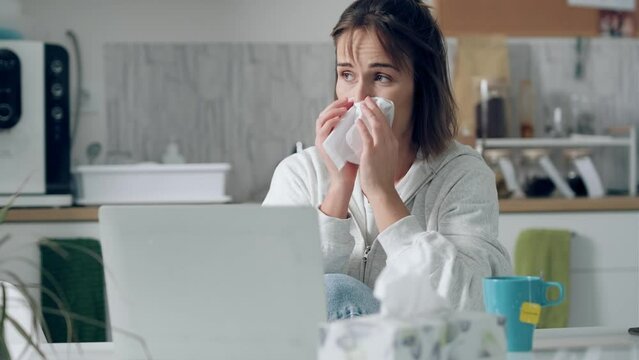 Video of llness young woman sneezing in a tissue while working with laptop sitting in the kitchen at home.