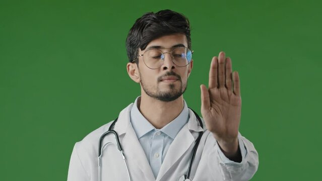 Young male doctor man in medical mask isolated in green chroma key studio show stop sign with prohibitive hand gesture safe distance puts palm forward disagreeing stop coronavirus infection spreading