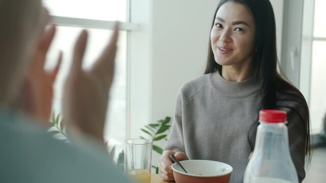 Happy Asian woman is eating cereal for breakfast and talking to husband smiling in kitchen at home. Conversation and healthy lifestyle concept.