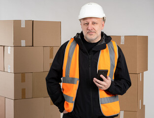 Warehouse manager. Man with phone in protective helmet. Warehouse manager in front of boxes. Warehouse manager uses mobile apps. Application for logistic accounting of concert. Guy in orange vest