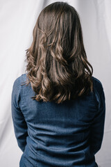 Woman photographed from the back showing her brunette hair. She is wearing a denim shirt against a white background