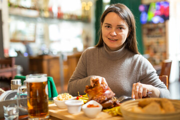 Positive brunette having lunch in Viennese cafe with traditional dish Schweinsstelze, crispy roasted ham hock with vegetable side dishes and glass of beer. Local cuisine - obrazy, fototapety, plakaty