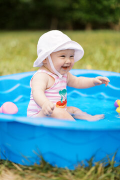 Adorable baby girl sits in a kiddy pool wearing a hat and swimsuit with chunky cheeks laughing. 