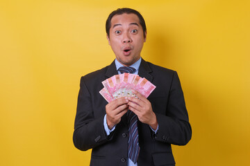 Asian businessman in suit standing against yellow background, showing money one hundred thousand rupiah on hands with wow expression. Financial and savings concept.