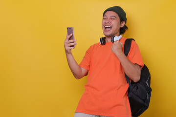 Attractive Asian young man in casual style with headphone and backpack is celebrating success against yellow background. Winning competition. Yes man. 