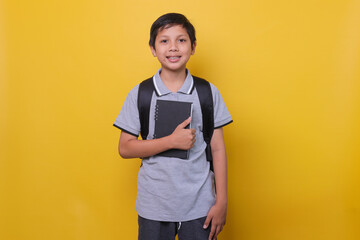 Asian school boy in casual style with black backpack is smiling and holding  a book isolated on...