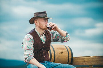 Cowboy drinks a whiskey on ranch. Handsome man in cowboy hat and retro vintage outfit.