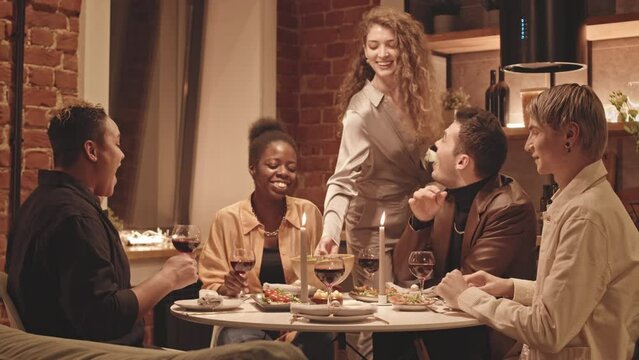 Medium slowmo of cheerful multiethnic gay and lesbian couple talking sitting at beautifully decorated dinner table while young woman with long curly hair serving dishes on table