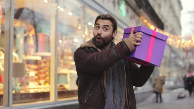 Excited bearded man in warm clothes shaking with gift box near ear while standing near christmas shop window. Caucasian guy guessing content of colorful present.