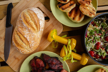 Dumbbells with tape measure, vegetable salad, whole loaf of bread, grilled pork neck meat from barbecue. Fit grill party concept, healthy or bad diet choice, fitness lifestyle gym workout composition.