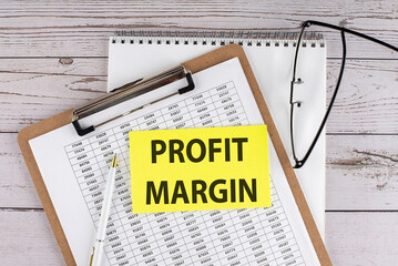 PROFIT MARGIN text on yellow sticky on clipboard with chart, wooden background