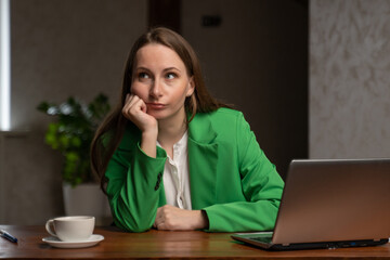 Young woman feels sad sitting at wooden table near laptop and cup of coffee. Bored brunette lady in elegant green jacket at workplace in office