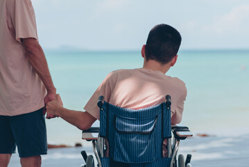 Wheelchair and behind of young man and father's, Dad hand holding the son's hand staring at the sea ahead,Family vacation activity,Encourage to strengthen,Mental health and travel or vacation concept. - obrazy, fototapety, plakaty