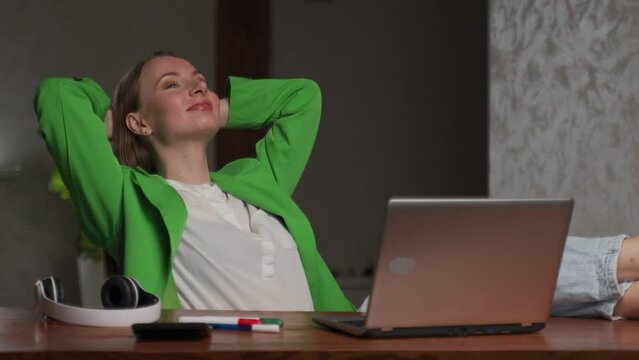 Young Woman In Stylish Green Jacket Takes Break From Work Leaning On Chair Back. Happy Lady Puts Hands Behind Head Smiling At Desk Near Open Laptop In Office