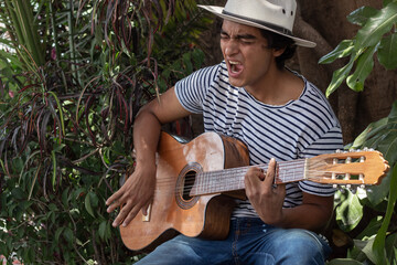 young hispanic guitarist with bohemian style playing his acoustic guitar outdoors in a park garden...