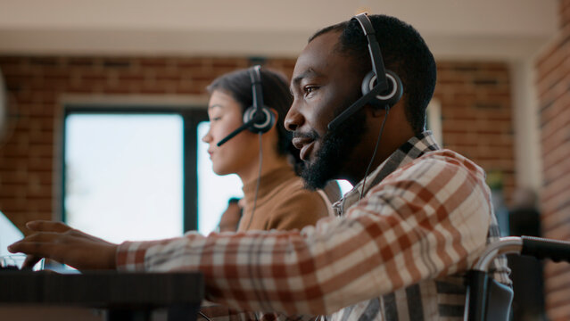 Male Worker Answering Phone Call On Audio Headset At Customer Service, Giving Help And Support In Office. Man In Wheelchair Using Headphones To Talk To People At Call Center. Close Up. Handheld Shot.