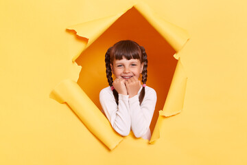 Portrait of adorable charming little girl with braids wearing casual shirt looking through torn hole in yellow paper, looking at camera with amazed eyes, keeps fists under chin.