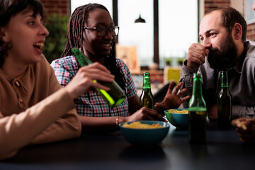 Friendly group of multicultural people at home talking while enjoying beer and snacks. Happy young adults sitting at table in living room while discussing and relaxing together.