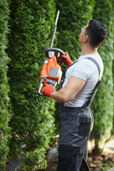 Naklejka premium Back view of professional gardener in uniform and gloves using hand electric trimmer for shaping hedge. Concept of seasonal work and landscaping. 
