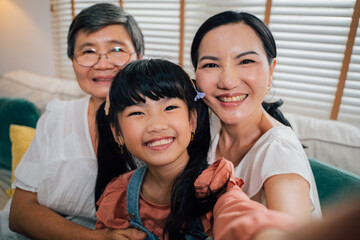 Cute Asian girl smiling and looking at camera while taking selfie with grandmother and aunt while...