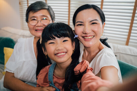 Cute Asian girl smiling and looking at camera while taking selfie with grandmother and aunt while sitting on sofa in living room at home