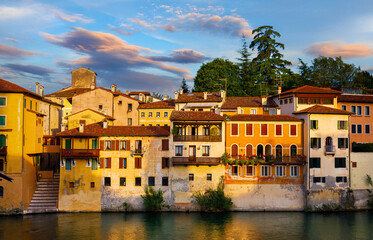 Fototapeta na wymiar Bassano del Grappa, Veneto, Italy. Ancient italian houses on river Brenta. Panoramic view at old town with vintage buildings of Alpine mountains scenic sunset landscape.