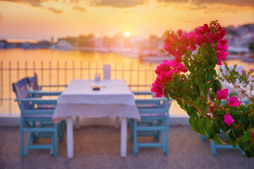 Sithonia, Chalkidiki, Greece. Traditional tavern cafe exterior with wooden table chairs. Flowers branch focused in front with the view of the aegean sea in the background. Sunset evening.