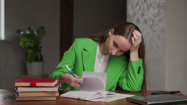 Tired teacher checks students homework sitting at table in classroom. Young woman in green jackets corrects mistakes in exercise books propping up head with hand