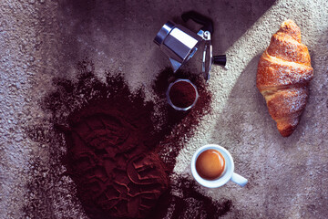 cup of italian espresso coffee bar with croissant and mocha full of ground coffee on gray stone surface and shoe print on coffee powder and grazing light