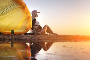 Attractive stylish young caucasian woman in cap sunglasses and kitesurfer outfit sits on the sandy shore next to her kite at sunset reflecting in the water - obrazy, fototapety, plakaty