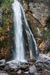 A girl in a green swimsuit sits under the streams of the waterfall. Grunas Waterfall is a picturesque site inside the National Park of Thethi, Albania