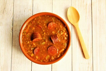 Clay bowl of lentils soup with Spanish sausage or chorizo with wooden spoon beside the bowl,...