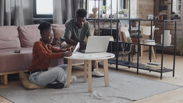 Long shot of happy Black hetero couple sitting in living room, planning family budget, using portable computer and having conversation
