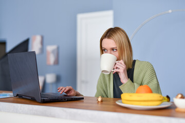 Young woman in the kitchen with mug and looking at her laptop