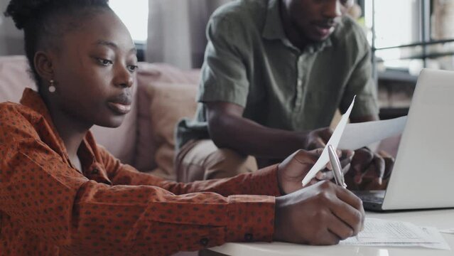 Medium close-up of young Black hetero couple sitting in living room, filling in tax returns, using portable computer and having conversation
