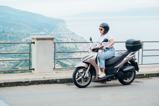 Middle-age woman in helmet and sunglasses on motor scooter on the Sicilian old town streets in the Forza d'Agro with Sant'Alessio Siculo harbor. Happy Italian vacation and transportation concept.