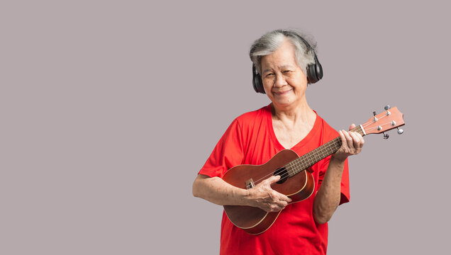 Portrait of an elderly Asian woman with short gray hair playing the ukulele with a smile while standing on a gray background