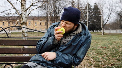 Hungry bearded homeless man sits on a bench and eats a green apple in a city park. Below poverty line. Male tramp in dirty clothes with cap and hat. Beggar. Illegal immigrant. Drunkenness, alcoholism