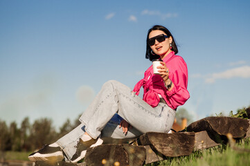 Brunette woman in sunglasses and pink shirt sitting and relaxing in the park over the blue sky background