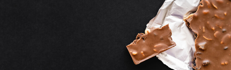 Top view of milk chocolate with nuts on foil on black background, banner.