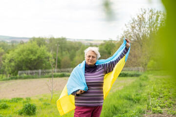 Senior white-haired woman wearing eyeglasses standing outdoors in the country waving the Ukrainian flag looking away feeling the freedom. No war, stop fights, we want peace.