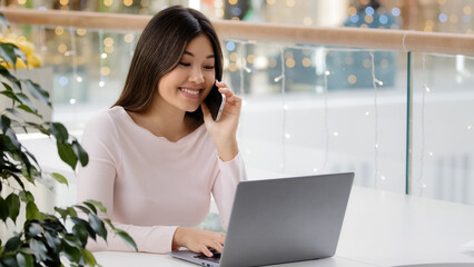 Happy Asian Korean woman freelancer student businesswoman holding mobile phone call working with laptop multitasking girl making online order communicating with friend distant friendly carefree talk