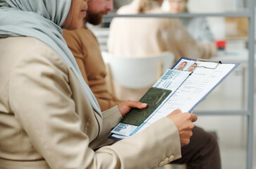Unrecognizable Muslim woman wearing hijab sitting on chair waiting for appointment in queue holding...