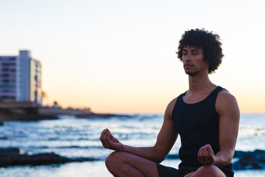 Young african american man with eyes closed meditating at beach during sunset, copy space