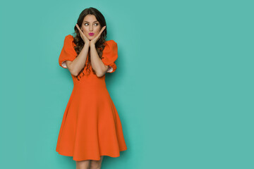 Beautiful young woman in orange mini dress is posing with head in hands and looking to the side.