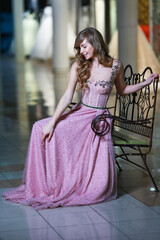 Fototapeta premium Beautiful blonde woman in a pink evening long dress sitting on a metal bench